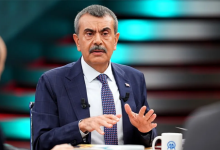 Bakan Yusuf Tekin, stüdyo ortamında koyu lacivert bir takım elbise, beyaz gömlek ve desenli bir kravatla oturuyor. Konuşurken elleriyle hafif jestler yapıyor. Ceketinde mikrofonlar takılı. Önünde AA logolu bir kupa ve bazı belgeler bulunuyor. Arka planda kırmızı ve turkuaz tonlarında aydınlatılmış stüdyo dekoru yer alıyor.