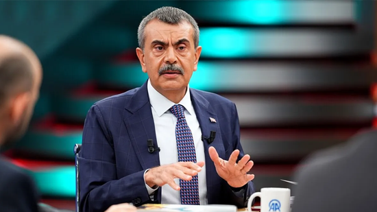 Bakan Yusuf Tekin, stüdyo ortamında koyu lacivert bir takım elbise, beyaz gömlek ve desenli bir kravatla oturuyor. Konuşurken elleriyle hafif jestler yapıyor. Ceketinde mikrofonlar takılı. Önünde AA logolu bir kupa ve bazı belgeler bulunuyor. Arka planda kırmızı ve turkuaz tonlarında aydınlatılmış stüdyo dekoru yer alıyor.