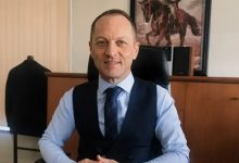 Murat Niyazoğlu, makam odasında çalışma masasında oturuyor. Üzerinde açık mavi bir gömlek, lacivert bir yelek ve desenli bir kravat bulunuyor. Ellerini masanın üzerinde birleştirmiş şekilde, sakin ve güven veren bir ifadeyle gülümsüyor. Arka planda ahşap bir dolap, üzerinde hoparlörler ve duvarda atlı bir figürün yer aldığı çerçeveli bir tablo görülüyor. Ortam resmi ve düzenli bir çalışma alanı izlenimi veriyor.