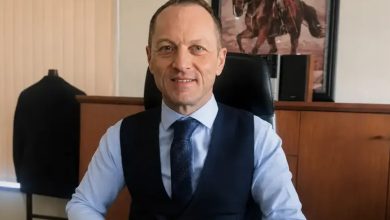 Murat Niyazoğlu, makam odasında çalışma masasında oturuyor. Üzerinde açık mavi bir gömlek, lacivert bir yelek ve desenli bir kravat bulunuyor. Ellerini masanın üzerinde birleştirmiş şekilde, sakin ve güven veren bir ifadeyle gülümsüyor. Arka planda ahşap bir dolap, üzerinde hoparlörler ve duvarda atlı bir figürün yer aldığı çerçeveli bir tablo görülüyor. Ortam resmi ve düzenli bir çalışma alanı izlenimi veriyor.