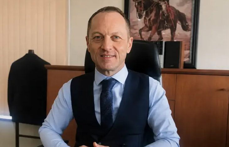 Murat Niyazoğlu, makam odasında çalışma masasında oturuyor. Üzerinde açık mavi bir gömlek, lacivert bir yelek ve desenli bir kravat bulunuyor. Ellerini masanın üzerinde birleştirmiş şekilde, sakin ve güven veren bir ifadeyle gülümsüyor. Arka planda ahşap bir dolap, üzerinde hoparlörler ve duvarda atlı bir figürün yer aldığı çerçeveli bir tablo görülüyor. Ortam resmi ve düzenli bir çalışma alanı izlenimi veriyor.