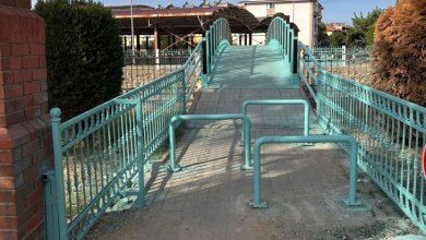 Görselin üst bölümünde, açık alanda bir su kanalının veya yolun üzerinden geçen açık yeşil boyalı kemerli yaya köprüsü ve köprüye bağlanan kilit taşlı yürüyüş yolu yer alıyor. Köprünün giriş kısmında tekerlekli sandalye veya bebek arabası geçişini zorlaştıracak şekilde ardışık olarak zemine monte edilmiş ters U biçiminde metal yeşil bariyerler bulunuyor. İki yanında dekoratif ferforje korkulukların uzandığı yolun çevresinde tuğla duvar, budanmış servi ağaçları, çalılar ve arka planda çok katlı yerleşim binaları görülüyor.