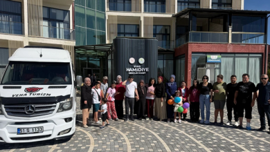 Görselin üst bölümünde, modern bir gençlik kampı tesisinin önündeki taş döşeli avluda bir araya gelmiş otizmli çocuklar, aileleri ve refakatçilerinden oluşan kalabalık bir grup yer alıyor. Sol tarafta "51 S 1133" plakalı ve üzerinde "VEHA TURİZM" yazan beyaz renkli bir Mercedes Sprinter minibüs park edilmiş durumda. Grubun hemen arkasında yer alan silindirik siyah giriş sütununda "SİVAS HAMİDİYE GENÇLİK KAMPI" yazısı ve resmi amblemler bulunuyor. Kadınlı erkekli, farklı yaşlardaki katılımcıların arasında elinde renkli balonlar tutan küçük bir kız çocuğu ile günlük yazlık kıyafetleriyle poz veren gençler ve aileler güler yüzle kameraya bakıyor; arka planda camlı ve balkonlu kamp binaları görülüyor.
