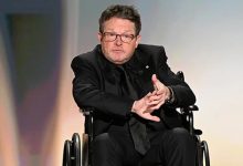 Michael J. Fox, tekerlekli sandalyede bulunuyor ve siyah takım elbiseli olarak konuşma yapıyor.