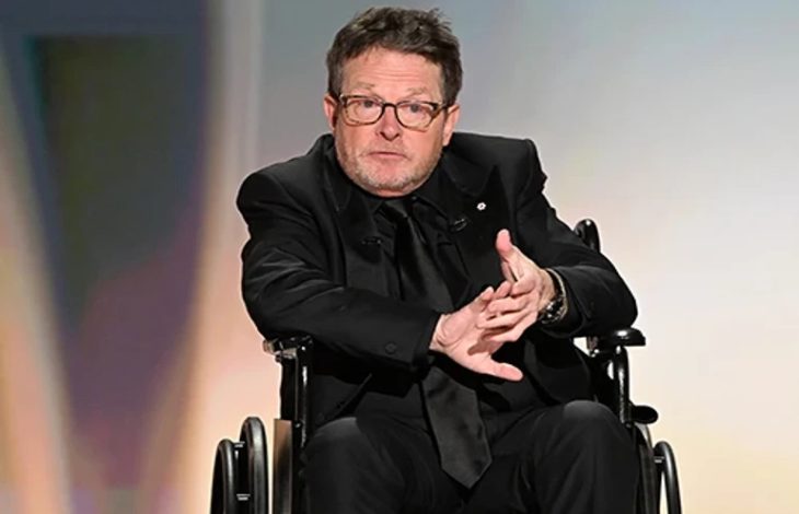 Michael J. Fox, tekerlekli sandalyede bulunuyor ve siyah takım elbiseli olarak konuşma yapıyor.