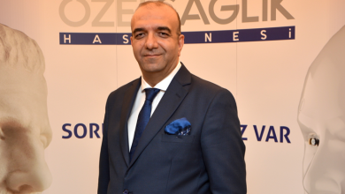 Görselde takım elbiseli Op. Dr. Kenan Kalı, üzerinde “Özel Sağlık Hastanesi” yazılı bir tanıtım panosunun önünde duruyor. Arka planda iki büyük heykel yüzü yer alıyor. Ortam resmi ve kurumsal bir sunum alanı izlenimi veriyor.