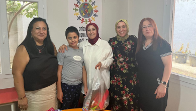Görselde, kırtasiye seti yardımı alan otizmli bir çocuk ailesiyle birlikte dernek yöneticilerinin yanında duruyor. Masanın üzerinde renkli kırtasiye malzemeleri bulunuyor. Yöneticilerden biri şeffaf bir poşet içinde eğitim materyallerini tutuyor. Arka planda otizme yönelik farkındalık afişi yer alıyor. Atmosfer sıcak, destekleyici ve dayanışma duygusunu yansıtıyor.