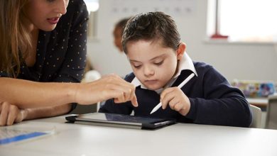 Sınıf ortamında, bir yetişkin ile bir çocuk yan yana oturuyor. Yetişkin tablet ekranını işaret ederken çocuk kalemle ekrana odaklanmış durumda. Arka planda sınıf eşyaları flu şekilde görülüyor.