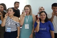 Bir grup kişi iç mekânda yan yana duruyor. Her biri elinde küçük bir kâğıt roket tutuyor ve roketi üfleyerek fırlatmaya hazırlanır gibi ağızlarına doğru götürmüş durumda. Üzerlerinde yaka kartları olan katılımcılar, etkinlik havasında neşeli ve dikkatli bir şekilde aynı hareketi yaparken görülüyor. Ortam aydınlık, samimi ve bir atölye ya da eğitim çalışması atmosferi taşıyor.