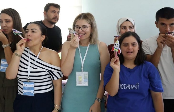Bir grup kişi iç mekânda yan yana duruyor. Her biri elinde küçük bir kâğıt roket tutuyor ve roketi üfleyerek fırlatmaya hazırlanır gibi ağızlarına doğru götürmüş durumda. Üzerlerinde yaka kartları olan katılımcılar, etkinlik havasında neşeli ve dikkatli bir şekilde aynı hareketi yaparken görülüyor. Ortam aydınlık, samimi ve bir atölye ya da eğitim çalışması atmosferi taşıyor.