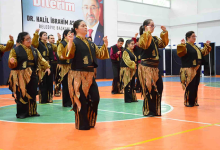 Fotoğrafta, bir spor salonunda sahne alan özel gereksinimli bireylerden oluşan geleneksel halk dansı ekibi görülüyor. Dansçılar, altın sarısı gömlekler, siyah yelekler ve siyah pantolonların üzerinde alev formunda altın detaylar bulunan uyumlu kostümler giymiş durumda. Spor salonunun zemini turuncu ve yeşil renkli bir saha çizimiyle kaplı. Arka planda büyük bir afiş yer alıyor. Afişte bir erkek portresi ve “DR. HALİL İBRAHİM ASLAN – BELEDİYE BAŞKANI” yazısı bulunuyor. Bu detay, etkinliğin bir belediye tarafından düzenlenen kültürel veya toplumsal bir organizasyon olduğunu düşündürüyor. Dansçılar senkronize bir şekilde poz verirken, ortamın genel atmosferi coşkulu ve geleneksel bir gösteri havası taşıyor.