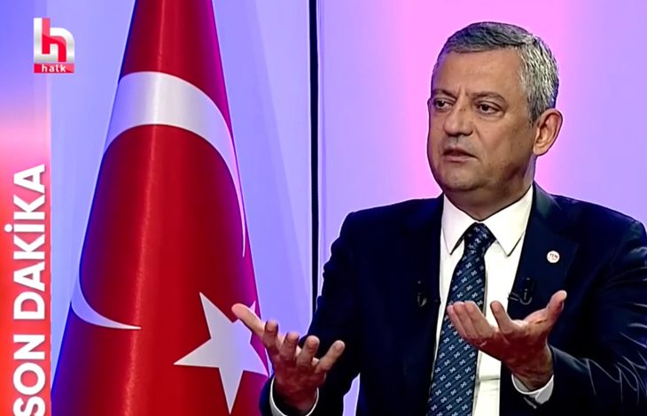 Stüdyoda takım elbise ve kravatıyla oturan Yeni Parti Genel Başkanı, canlı yayında konuşurken iki elini hafifçe yukarı doğru açmış şekilde jest yapıyor. Arka planda mor ve pembe tonlarda aydınlatılmış bir stüdyo atmosferi bulunuyor. Sağ tarafta büyük bir Türk bayrağı yer alıyor. Sol tarafta kırmızı zemin üzerinde beyaz harflerle “SON DAKİKA” yazısı ve onun üstünde kanalın logosu görünüyor. Genel başkan, ciddi ve odaklanmış bir ifadeyle gündeme dair açıklama yapıyor gibi görünüyor.