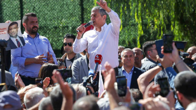 Özgür Özel, açık alanda kalabalığa hitap ederken görülüyor. Bir kürsü veya mikrofon önünde konuşma yapıyor; etrafında partililer ve vatandaşlar bulunuyor. Özel, el hareketleri ve yüz ifadesiyle vurgulu bir konuşma yaptığı izlenimi veriyor. Arkada parti bayrakları veya etkinlik alanına ait görseller yer alıyor.
