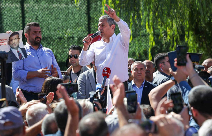 Özgür Özel, açık alanda kalabalığa hitap ederken görülüyor. Bir kürsü veya mikrofon önünde konuşma yapıyor; etrafında partililer ve vatandaşlar bulunuyor. Özel, el hareketleri ve yüz ifadesiyle vurgulu bir konuşma yaptığı izlenimi veriyor. Arkada parti bayrakları veya etkinlik alanına ait görseller yer alıyor.