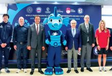 Yedi kişi, “World Para Swimming European Championships Kocaeli 2026” yazılı tanıtım panosu önünde poz veriyor. Ortada mavi, sevimli bir su canlısı maskotu duruyor. Ortam resmi bir tanıtım etkinliği havasında.