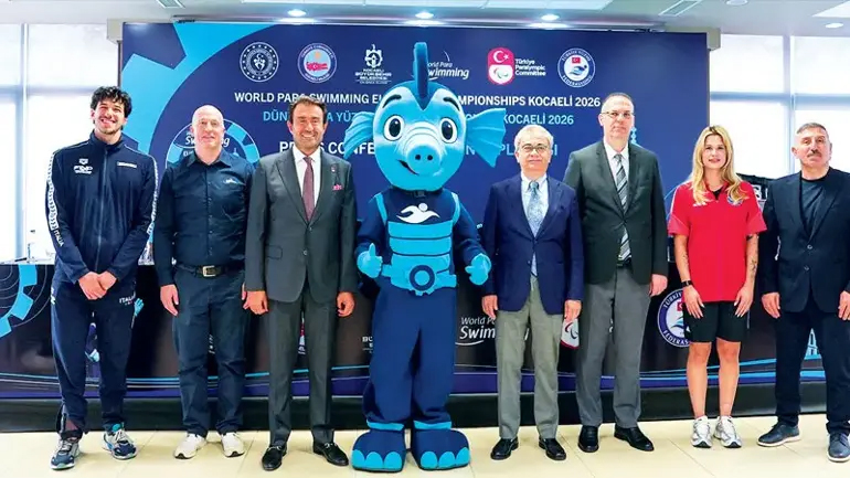 Yedi kişi, “World Para Swimming European Championships Kocaeli 2026” yazılı tanıtım panosu önünde poz veriyor. Ortada mavi, sevimli bir su canlısı maskotu duruyor. Ortam resmi bir tanıtım etkinliği havasında.