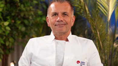 Prof. Dr. Burak Turna, beyaz bir tıbbi önlük ile gri bir koltukta oturuyor. Önlüğün sol göğüs kısmında “American Hastanesi” yazısı ve altında “Doctor” ibaresi yer alıyor. Arka planda yeşil bitkiler ve palm yaprakları bulunuyor; ortamın ışığı doktora sıcak bir ton verirken arka plan daha mavi ve loş bir görünüm taşıyor. Doktor sakin, kendinden emin ve profesyonel bir duruşla kameraya doğru bakıyor.