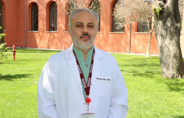 Açık havada, bakımlı bir çim alan üzerinde ayakta duran bir erkek görülüyor. Üzerinde beyaz hekim önlüğü, altında yeşil bir üst ve boynunda bordo renkli, üzerinde yazılar bulunan bir yaka kartı askısı bulunuyor. Önlüğün üzerinde “PROF. DR. ÜNAL AKIN” yazılı bir isim etiketi yer alıyor. Yaka kartı kırmızı bir klipsle tutturulmuş. Arka planda turuncu tonlarda bir bina, kemerli pencereler, seyrek yapraklı ağaçlar ve çim alanda bir yangın musluğu görülüyor. Duruşu ve ortam, kendisini tıbbi uzmanlığı güçlü, akademik kimliği belirgin ve profesyonel bir hekim olarak yansıtıyor.