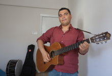Görselin üst bölümünde, sade beyaz duvarlı bir odada ayakta durarak klasik gitar çalan genç bir erkek yer alıyor. Bordo renkli uzun kollu gömlek ve kot pantolon giymiş olan otizmli müzisyen, boynuna astığı gitar askısıyla açık ahşap renkli klasik gitarını çalarken hafifçe gülümseyerek doğrudan kameraya bakıyor. Sol arka planda ise zeminde duran siyah bir davul ile duvara yaslanmış siyah bir enstrüman kılıfı (gitar çantası) görülüyor.