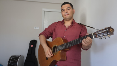 Görselin üst bölümünde, sade beyaz duvarlı bir odada ayakta durarak klasik gitar çalan genç bir erkek yer alıyor. Bordo renkli uzun kollu gömlek ve kot pantolon giymiş olan otizmli müzisyen, boynuna astığı gitar askısıyla açık ahşap renkli klasik gitarını çalarken hafifçe gülümseyerek doğrudan kameraya bakıyor. Sol arka planda ise zeminde duran siyah bir davul ile duvara yaslanmış siyah bir enstrüman kılıfı (gitar çantası) görülüyor.