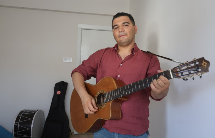 Görselin üst bölümünde, sade beyaz duvarlı bir odada ayakta durarak klasik gitar çalan genç bir erkek yer alıyor. Bordo renkli uzun kollu gömlek ve kot pantolon giymiş olan otizmli müzisyen, boynuna astığı gitar askısıyla açık ahşap renkli klasik gitarını çalarken hafifçe gülümseyerek doğrudan kameraya bakıyor. Sol arka planda ise zeminde duran siyah bir davul ile duvara yaslanmış siyah bir enstrüman kılıfı (gitar çantası) görülüyor.