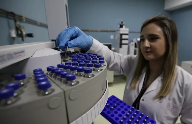 Bir sağlık çalışanı laboratuvarda beyaz önlük ve mavi eldivenlerle küçük numune tüplerini dikkatle yerleştiriyor. Önünde dairesel bir örnek tepsisi ve analiz cihazı bulunuyor; ortam steril ve profesyonel bir çalışma alanı hissi veriyor.