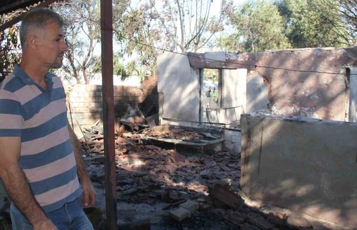 Yangın sonrası ev tamamen harap durumda. Duvarlar çökmüş, tuğlalar ve beton parçaları etrafa dağılmış. Çatı yok, iç kısımlar açıkta. Önde bir kişi hasarı inceliyor.