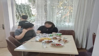Anne ve çocuklarından biri yemek masasında oturuyor, diğer çocuğu ise camdan dışarı bakıyor.