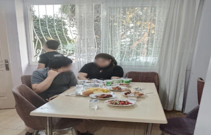 Anne ve çocuklarından biri yemek masasında oturuyor, diğer çocuğu ise camdan dışarı bakıyor.