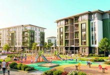 Modern bir sosyal konut alanı görülüyor. Orta katlı, yeşil ve beyaz tonlarda apartmanlar siteyi çevreliyor. Ortada renkli bir çocuk oyun parkı var; kaydıraklar, salıncaklar ve tırmanma alanları bulunuyor. Çocuklar oynarken yetişkinler çevrede yürüyüş yapıyor veya onları izliyor. Alan temiz, düzenli ve yeşilliklerle çevrili.