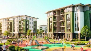 Modern bir sosyal konut alanı görülüyor. Orta katlı, yeşil ve beyaz tonlarda apartmanlar siteyi çevreliyor. Ortada renkli bir çocuk oyun parkı var; kaydıraklar, salıncaklar ve tırmanma alanları bulunuyor. Çocuklar oynarken yetişkinler çevrede yürüyüş yapıyor veya onları izliyor. Alan temiz, düzenli ve yeşilliklerle çevrili.
