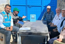 Görselin üst bölümünde, açık alanda mavi bir demir kapının önündeki masa etrafında oturarak sohbet eden bir heyet ve vatandaşlar yer alıyor. Sol tarafta mavi belediye yeleği, beyaz gömlek ve kravat giymiş bir görevli ile hemen yanında yeşil şapkalı tekerlekli sandalye kullanan engelli bir birey oturuyor. Çizgili masa örtüsü serili masanın üzerinde not panosu, kalem ve üzerinde Talas Belediyesi amblemi bulunan açık mavi bir hediye çantası duruyor. Masanın sağ tarafında ise kasketli, ekose gömlekli ve beyaz gömlekli yaşlı mahalle sakinleri ile aile bireyleri yan yana oturarak ziyarete eşlik ediyor.