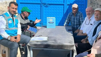 Görselin üst bölümünde, açık alanda mavi bir demir kapının önündeki masa etrafında oturarak sohbet eden bir heyet ve vatandaşlar yer alıyor. Sol tarafta mavi belediye yeleği, beyaz gömlek ve kravat giymiş bir görevli ile hemen yanında yeşil şapkalı tekerlekli sandalye kullanan engelli bir birey oturuyor. Çizgili masa örtüsü serili masanın üzerinde not panosu, kalem ve üzerinde Talas Belediyesi amblemi bulunan açık mavi bir hediye çantası duruyor. Masanın sağ tarafında ise kasketli, ekose gömlekli ve beyaz gömlekli yaşlı mahalle sakinleri ile aile bireyleri yan yana oturarak ziyarete eşlik ediyor.