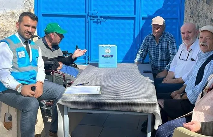 Görselin üst bölümünde, açık alanda mavi bir demir kapının önündeki masa etrafında oturarak sohbet eden bir heyet ve vatandaşlar yer alıyor. Sol tarafta mavi belediye yeleği, beyaz gömlek ve kravat giymiş bir görevli ile hemen yanında yeşil şapkalı tekerlekli sandalye kullanan engelli bir birey oturuyor. Çizgili masa örtüsü serili masanın üzerinde not panosu, kalem ve üzerinde Talas Belediyesi amblemi bulunan açık mavi bir hediye çantası duruyor. Masanın sağ tarafında ise kasketli, ekose gömlekli ve beyaz gömlekli yaşlı mahalle sakinleri ile aile bireyleri yan yana oturarak ziyarete eşlik ediyor.