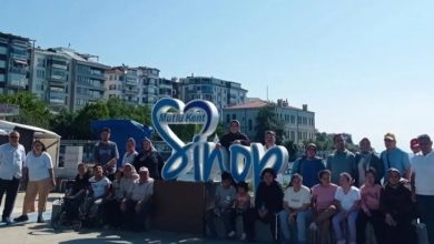 Görselde, “Mutlu Kent Sinop” yazılı büyük dekoratif yapının önünde bir grup insan dış mekânda toplu hâlde poz veriyor. Arkada apartmanlar ve ağaçlar görülüyor. Hava güneşli; ortam samimi, şehir meydanında çekilmiş bir hatıra fotoğrafı izlenimi veriyor.