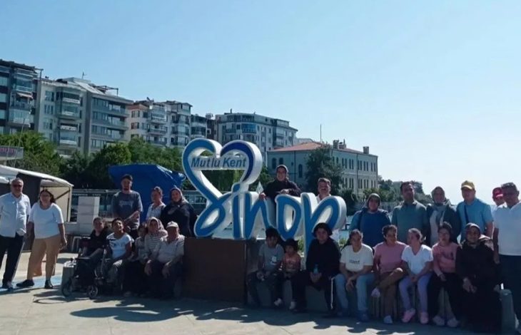 Görselde, “Mutlu Kent Sinop” yazılı büyük dekoratif yapının önünde bir grup insan dış mekânda toplu hâlde poz veriyor. Arkada apartmanlar ve ağaçlar görülüyor. Hava güneşli; ortam samimi, şehir meydanında çekilmiş bir hatıra fotoğrafı izlenimi veriyor.