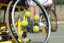Sarı çerçeveli bir tekerlekli sandalye tenis kortunda duruyor. Tekerleğin tellerine birkaç tenis topu sıkıştırılmış. Arka planda başka bir tekerlekli sandalye daha görülüyor; sahne engelli sporlarına yönelik bir tenis antrenmanını çağrıştırıyor.