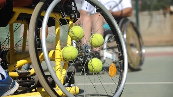 Sarı çerçeveli bir tekerlekli sandalye tenis kortunda duruyor. Tekerleğin tellerine birkaç tenis topu sıkıştırılmış. Arka planda başka bir tekerlekli sandalye daha görülüyor; sahne engelli sporlarına yönelik bir tenis antrenmanını çağrıştırıyor.