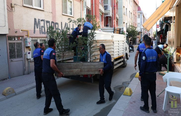 Görselde, dar bir sokakta belediye zabıta ekiplerinin bir kamyonete yükledikleri uzun ahşap masayı taşırken ve çevredeki eşyaları düzenlerken görülüyor.
