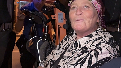 Görselin üst bölümünde, bir polis aracının veya resmi otomobilin içinde oturan orta yaşlı bir anne ve arkada ona yardım eden polis memuru yer alıyor. Ön koltukta oturan, başında çiçek desenli renkli bir tülbent ve üzerinde siyah-beyaz desenli yazlık gömlek bulunan anne, doğrudan merceğe bakarak hafifçe konuşur gibi duruyor. Aracın arka kısmında ise mavi üniformalı bir polis memuru, engelli bireyin akülü sandalyesini veya tekerlekli aparatını araca yerleştirmek için özenle ilgileniyor.