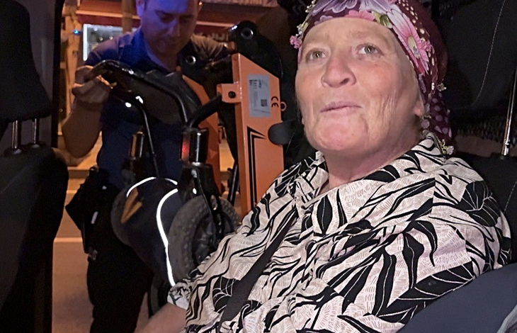 Görselin üst bölümünde, bir polis aracının veya resmi otomobilin içinde oturan orta yaşlı bir anne ve arkada ona yardım eden polis memuru yer alıyor. Ön koltukta oturan, başında çiçek desenli renkli bir tülbent ve üzerinde siyah-beyaz desenli yazlık gömlek bulunan anne, doğrudan merceğe bakarak hafifçe konuşur gibi duruyor. Aracın arka kısmında ise mavi üniformalı bir polis memuru, engelli bireyin akülü sandalyesini veya tekerlekli aparatını araca yerleştirmek için özenle ilgileniyor.