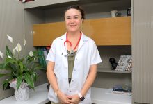 Uzm. Dr. Dilek Dalkıran Erdem, beyaz hekim önlüğü ve boynunda kırmızı stetoskopla ayakta duran bir sağlık profesyoneli olarak görülüyor. Arka planda kitaplık, dekoratif objeler ve çiçek bulunan sade bir klinik/ofis ortamı yer alıyor. Duruşu sakin ve güven veren bir izlenim oluşturuyor.