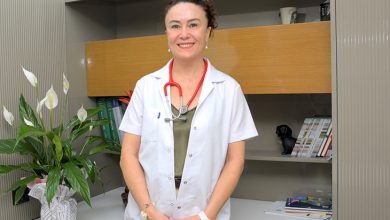 Uzm. Dr. Dilek Dalkıran Erdem, beyaz hekim önlüğü ve boynunda kırmızı stetoskopla ayakta duran bir sağlık profesyoneli olarak görülüyor. Arka planda kitaplık, dekoratif objeler ve çiçek bulunan sade bir klinik/ofis ortamı yer alıyor. Duruşu sakin ve güven veren bir izlenim oluşturuyor.