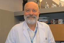 Fotoğrafta Uzm. Dr. Atilla Kara, beyaz bir doktor önlüğü ve boynunda mavi bir yaka kartı ile görülüyor. Sakallı, orta yaş üstü bir görünümü var ve kameraya doğru sakin bir ifadeyle bakıyor. Arka planda raflar bulunuyor; raflarda kitaplar, kupalar ve küçük dekoratif objeler yer alıyor. Kitaplardan biri “Bu Da Geçer” başlığını taşıyor. Ortam, bir doktorun çalışma odası ya da danışanlarını karşıladığı profesyonel bir ofis izlenimi veriyor.