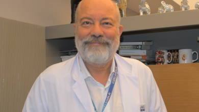 Fotoğrafta Uzm. Dr. Atilla Kara, beyaz bir doktor önlüğü ve boynunda mavi bir yaka kartı ile görülüyor. Sakallı, orta yaş üstü bir görünümü var ve kameraya doğru sakin bir ifadeyle bakıyor. Arka planda raflar bulunuyor; raflarda kitaplar, kupalar ve küçük dekoratif objeler yer alıyor. Kitaplardan biri “Bu Da Geçer” başlığını taşıyor. Ortam, bir doktorun çalışma odası ya da danışanlarını karşıladığı profesyonel bir ofis izlenimi veriyor.