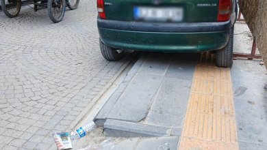 Görselin üst bölümünde, kaldırım üzerine park ederek geçişi engelleyen koyu yeşil bir binek otomobil yer alıyor. Plakası buzlanmış olan aracın sağ tekerlekleri doğrudan kaldırım üzerindeki sarı kabartmalı (hissedilebilir) görme engelli yürüyüş bandının üzerinde dururken, sol tekerlekleri tekerlekli sandalye geçişi için ayrılmış eğimli rampa kısmını tamamen kapatıyor. Sol tarafta kilit taşlı yolda duran mavi gövdeli seyyar bir arabanın tekerlekleri, ön planda ise kaldırım kenarına atılmış boş su şişesi ve plastik çöp poşeti görülüyor.