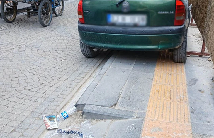 Görselin üst bölümünde, kaldırım üzerine park ederek geçişi engelleyen koyu yeşil bir binek otomobil yer alıyor. Plakası buzlanmış olan aracın sağ tekerlekleri doğrudan kaldırım üzerindeki sarı kabartmalı (hissedilebilir) görme engelli yürüyüş bandının üzerinde dururken, sol tekerlekleri tekerlekli sandalye geçişi için ayrılmış eğimli rampa kısmını tamamen kapatıyor. Sol tarafta kilit taşlı yolda duran mavi gövdeli seyyar bir arabanın tekerlekleri, ön planda ise kaldırım kenarına atılmış boş su şişesi ve plastik çöp poşeti görülüyor.