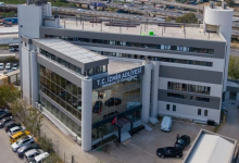 Görselde, modern mimariye sahip çok katlı Yeni İzmir Adliyesi görülüyor. Bina geniş cam cepheli, gri tonlarda tasarlanmış ve giriş bölümünde silindirik kolonlar bulunuyor. Üst kısımda “T.C. İzmir Adliyesi – Halkapınar Ek Hizmet Binası” yazısı yer alıyor. Ön tarafta park halinde araçlar ve girişe yönelen birkaç kişi görülüyor. Arka planda yol, araç trafiği ve tren hatları yer alarak yapının kent içindeki konumunu gösteriyor. Bina, yeni ve çağdaş bir kamu hizmet yapısı izlenimi veriyor.