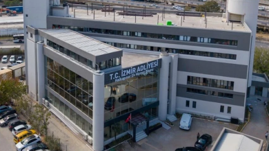 Görselde, modern mimariye sahip çok katlı Yeni İzmir Adliyesi görülüyor. Bina geniş cam cepheli, gri tonlarda tasarlanmış ve giriş bölümünde silindirik kolonlar bulunuyor. Üst kısımda “T.C. İzmir Adliyesi – Halkapınar Ek Hizmet Binası” yazısı yer alıyor. Ön tarafta park halinde araçlar ve girişe yönelen birkaç kişi görülüyor. Arka planda yol, araç trafiği ve tren hatları yer alarak yapının kent içindeki konumunu gösteriyor. Bina, yeni ve çağdaş bir kamu hizmet yapısı izlenimi veriyor.