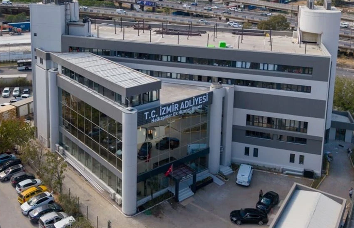 Görselde, modern mimariye sahip çok katlı Yeni İzmir Adliyesi görülüyor. Bina geniş cam cepheli, gri tonlarda tasarlanmış ve giriş bölümünde silindirik kolonlar bulunuyor. Üst kısımda “T.C. İzmir Adliyesi – Halkapınar Ek Hizmet Binası” yazısı yer alıyor. Ön tarafta park halinde araçlar ve girişe yönelen birkaç kişi görülüyor. Arka planda yol, araç trafiği ve tren hatları yer alarak yapının kent içindeki konumunu gösteriyor. Bina, yeni ve çağdaş bir kamu hizmet yapısı izlenimi veriyor.